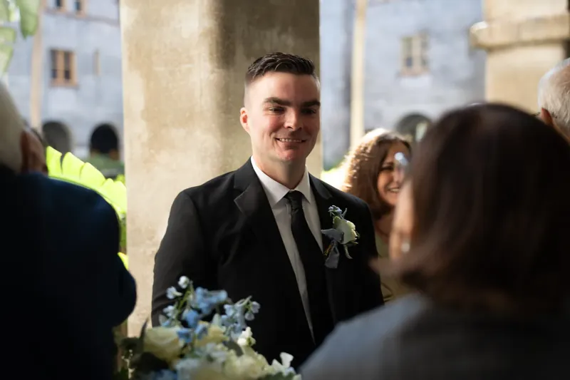 Bailey stands in the courtyard at Lightner Museum wearing a dark suit and boutonniere, greeting guests during the reception.