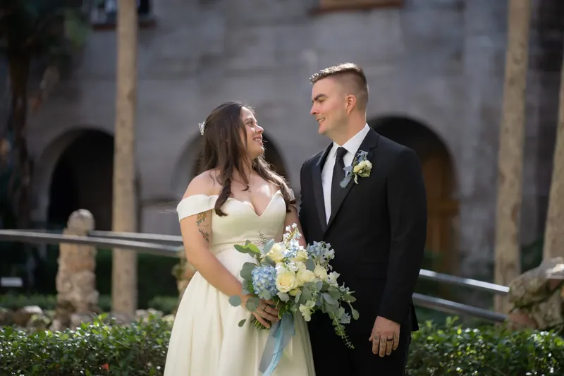 Bailey gazes at Ana while standing beside hedges at the Lightner Museum, both dressed formally for their ceremony.