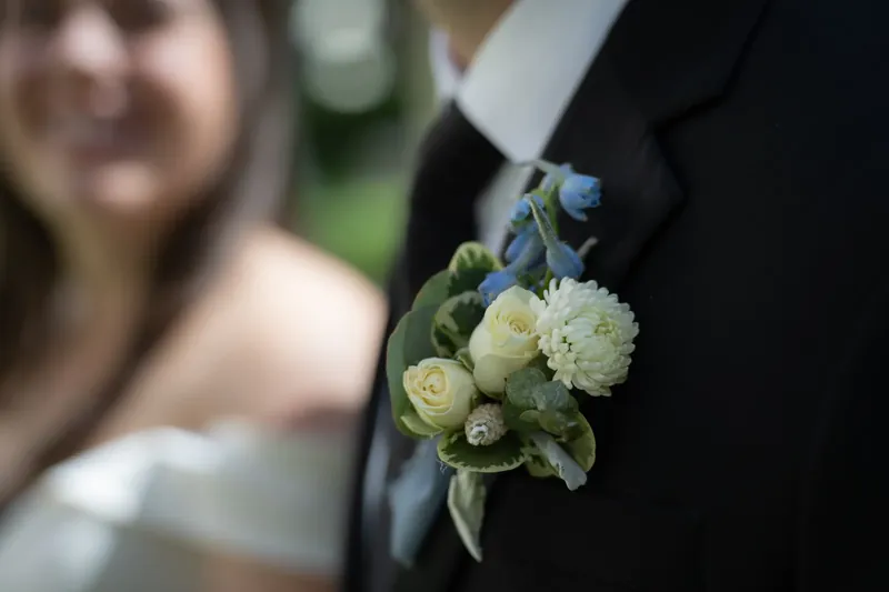 A white and blue boutonniere pinned to Bailey's dark suit jacket, with Ana visible softly in the background.