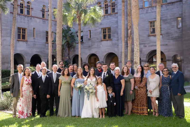 The entire wedding party stands together on the lawn in front of the Lightner Museum's arched stone facade and palm trees.