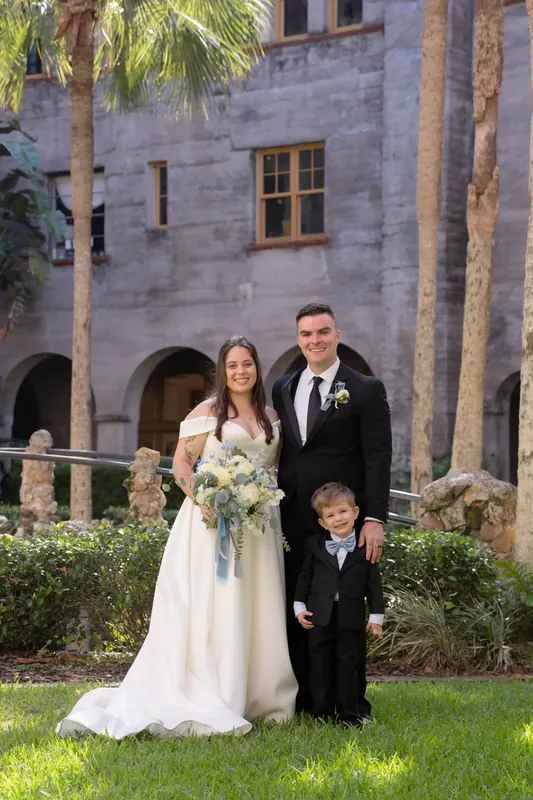 Ana, Bailey, and a young child in formal wear stand on the grass with the historic courtyard archways behind them.
