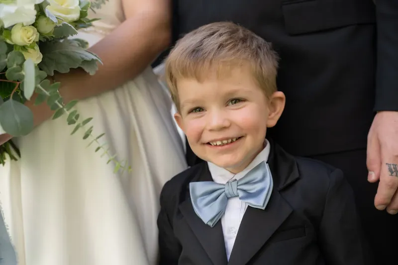 A young ring bearer in a tuxedo and light blue bow tie grins at the camera beside the bride's bouquet.