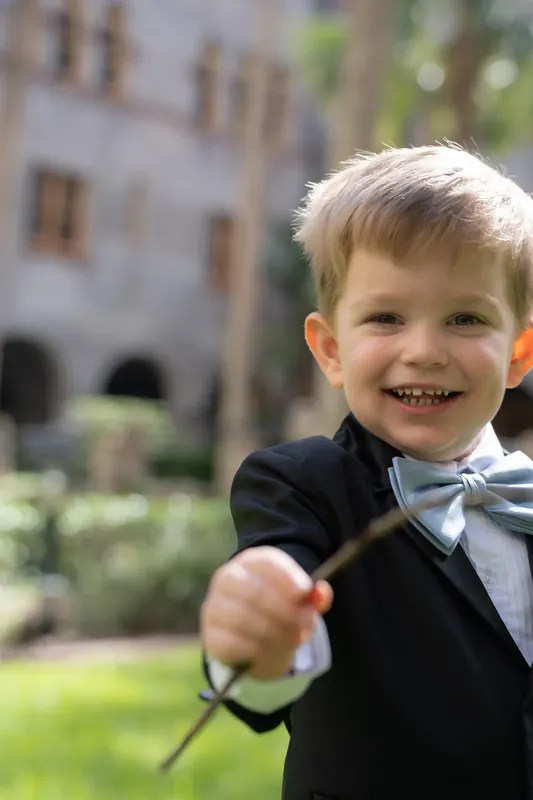 A young attendant in a black tuxedo and bow tie holds the ring pillow while beaming at the camera.