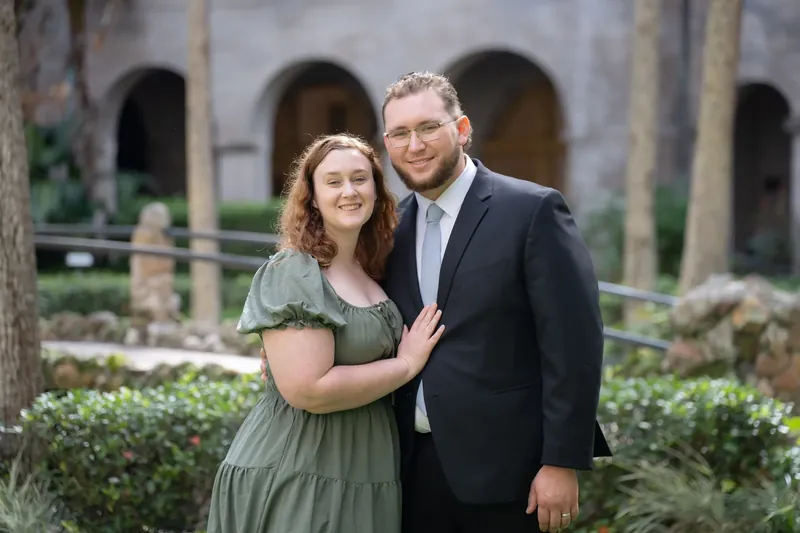 Ana and Bailey embrace in the Lightner Museum courtyard, arched stone architecture and trimmed hedges behind them.
