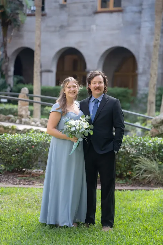 Bailey holds a white bouquet while posing with Ana on the museum's lawn, stone archways visible in the background.