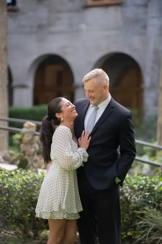 Ana in a cream polka-dot dress looks up at Bailey as they stand near the columned courtyard of the historic venue.