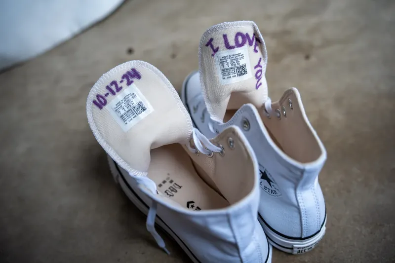 White sneakers customized with purple handwriting rest on stone flooring, reading "I do" and "Love.