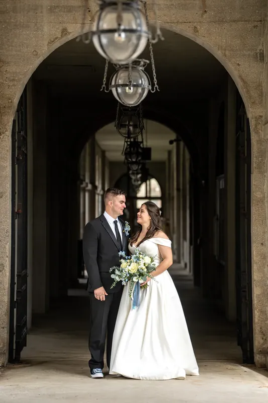 Ana and Bailey face each other beneath a stone archway inside Lightner Museum, holding their wedding flowers.