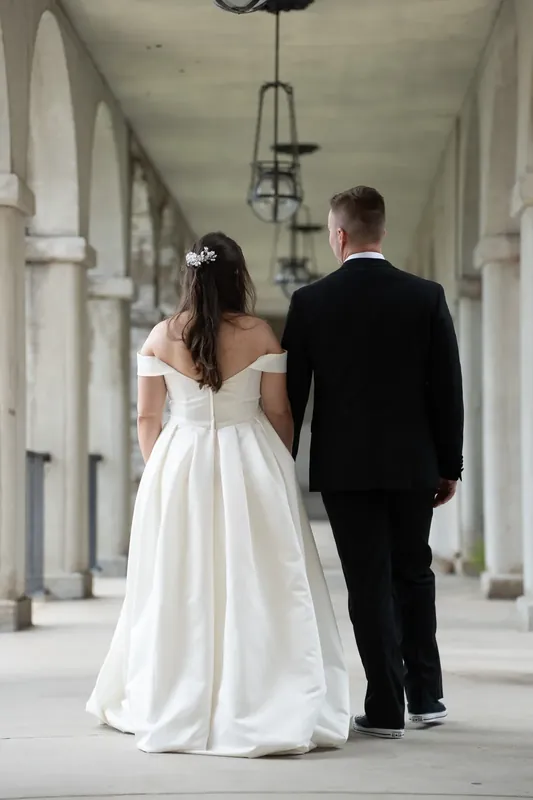 Ana and Bailey walk hand-in-hand down the columned corridor at Lightner Museum in their wedding attire.