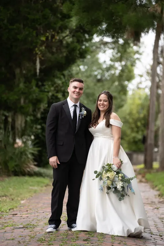 Ana and Bailey stand together on a tree-lined path, Ana holding a greenery bouquet and smiling at the camera.