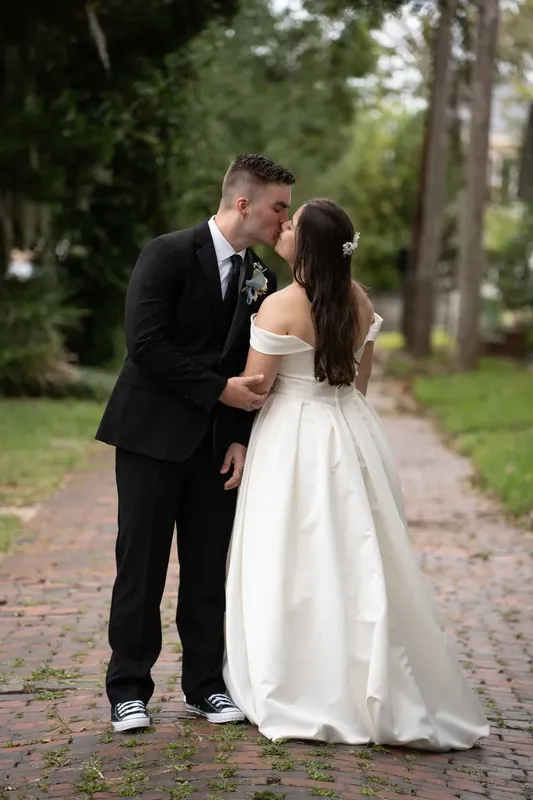 Bailey kisses Ana's cheek as they stand on a brick pathway lined with oak trees at Lightner Museum.