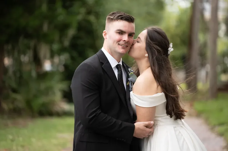 Ana leans in to kiss Bailey's cheek while standing outdoors under the canopy of trees at Lightner Museum.