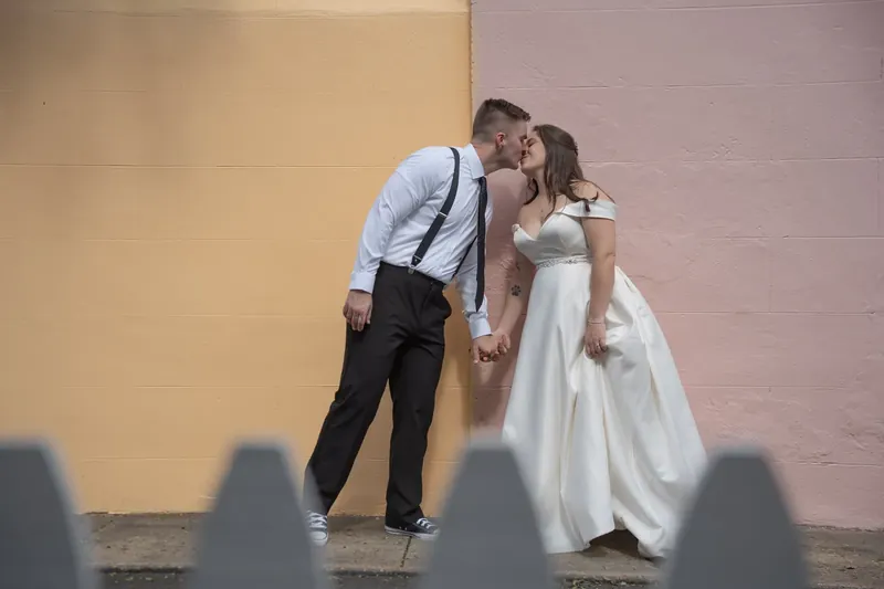 Bailey leans in to kiss Ana beneath an archway, their shadows cast across the pink and yellow walls behind them.