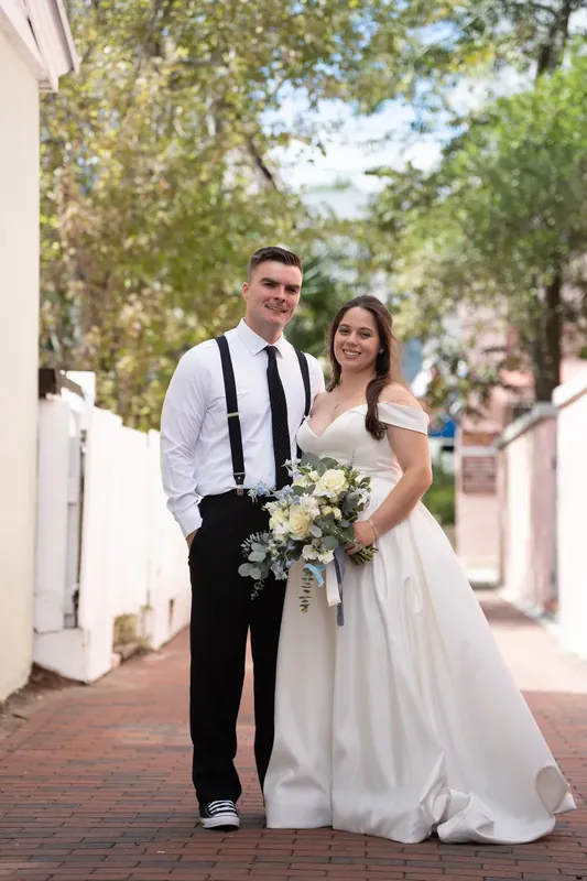 Ana and Bailey walk a brick pathway at Lightner Museum, Ana holding a white bouquet in a flowing gown while Bailey wears a black tie and suspenders.