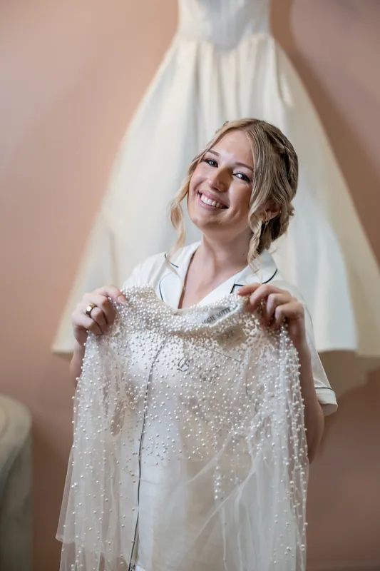 Anna holds up a beaded wedding cape and smiles while standing before her white wedding dress at The Treasury on the Plaza