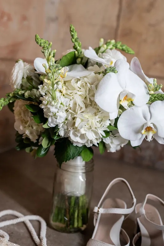Bridal bouquet of white orchids, cream hydrangeas, and green foliage in a clear vase sits beside white wedding heels.