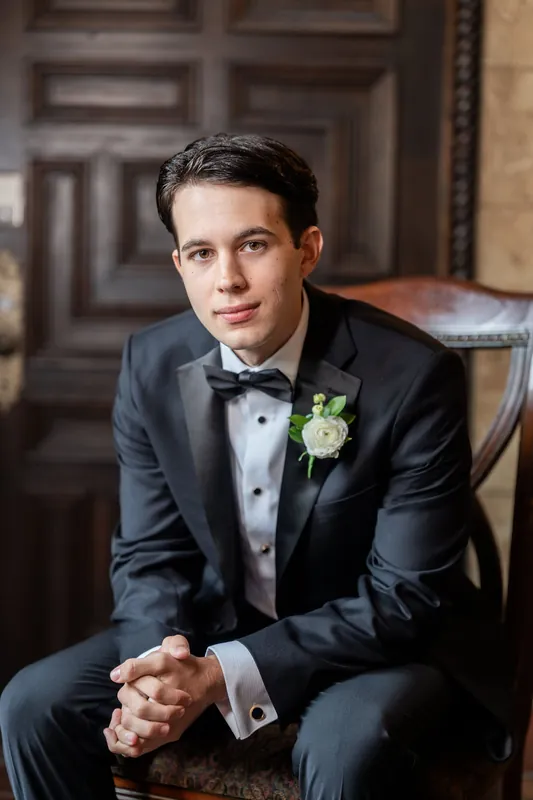 Rafael sits in a dark tuxedo with a white boutonniere, hands clasped, in an ornate interior hallway.
