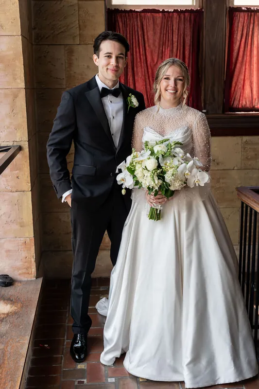 Anna carries a white floral bouquet alongside Rafael in his tuxedo as they walk through the stone archway.