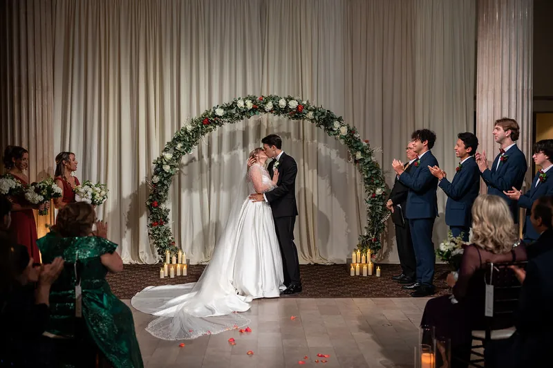 Anna and Rafael exchange vows beneath a greenery arch with their wedding party standing nearby.