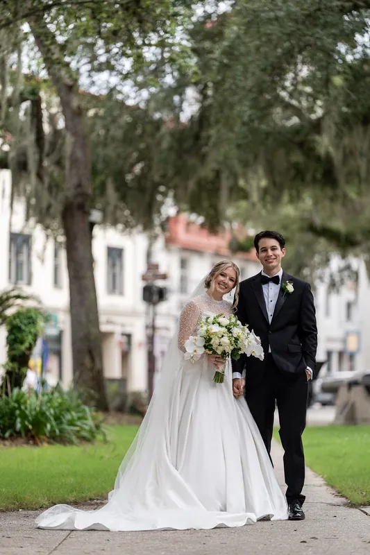 Anna and Rafael walk arm-in-arm beneath live oaks at The Treasury on the Plaza, Anna holding a white bouquet in a flowing