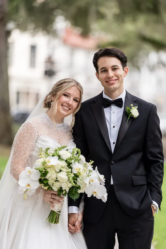 Rafael smiles beside Anna, who holds a lush white and green bouquet, both dressed formally in front of historic brick