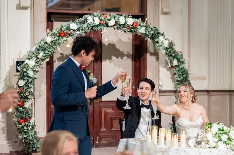 A groomsman delivers a toast while Anna, Rafael, and a guest listen at their reception dinner table.