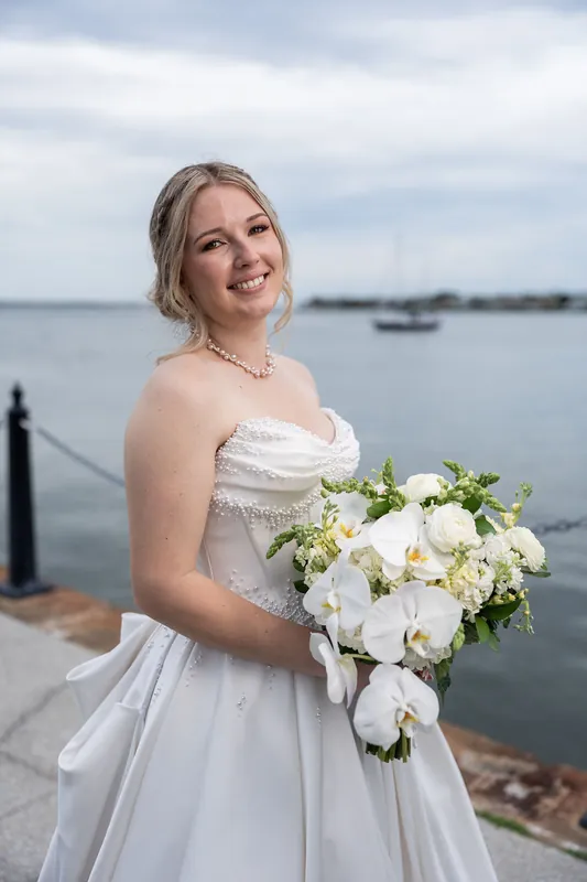 Anna smiles at the camera holding her white and green bouquet with a waterfront and boats behind her.