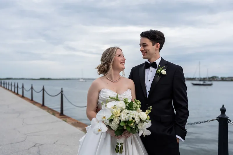 Rafael and Anna walk together along a waterfront railing with boats visible across the water behind them.