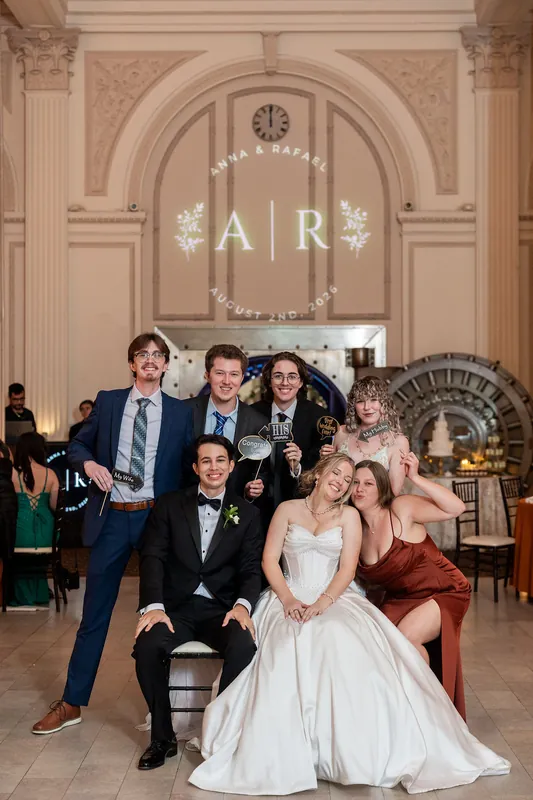 Anna in a strapless white gown and Rafael in a black tuxedo pose with family members beneath an ornate archway decorated
