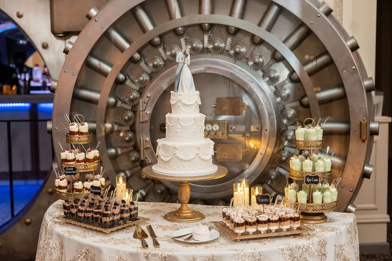 A three-tiered white wedding cake sits on a gold stand surrounded by desserts and candles, framed by an elaborate spiral