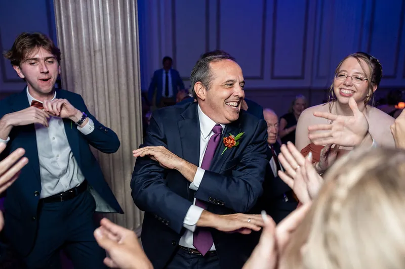 A man in a navy suit and pink boutonniere dances enthusiastically with other guests on the reception floor.