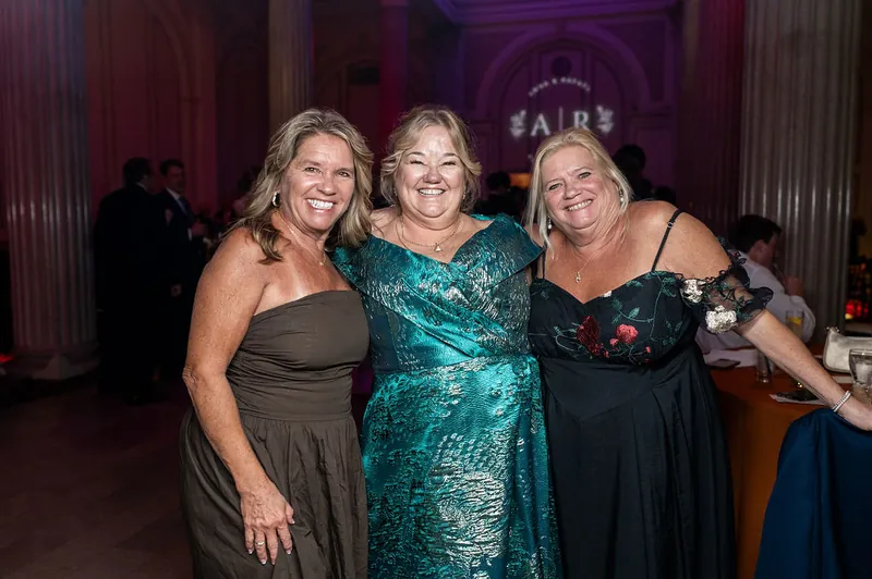 Three women smile together at the reception, dressed in formal evening wear with the venue's grand interior visible behind