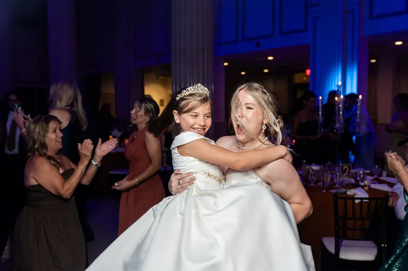 A child in a white dress hugs a woman in a white gown while guests applaud at The Treasury on the Plaza, St. Augustine.