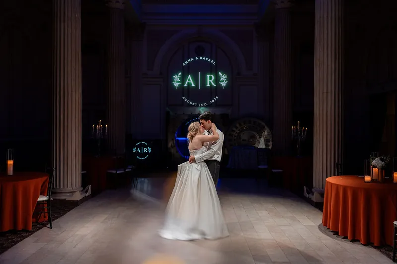 Anna and Rafael kiss under glowing "AIR" initials in a columned hall lined with orange-draped tables at The Treasury