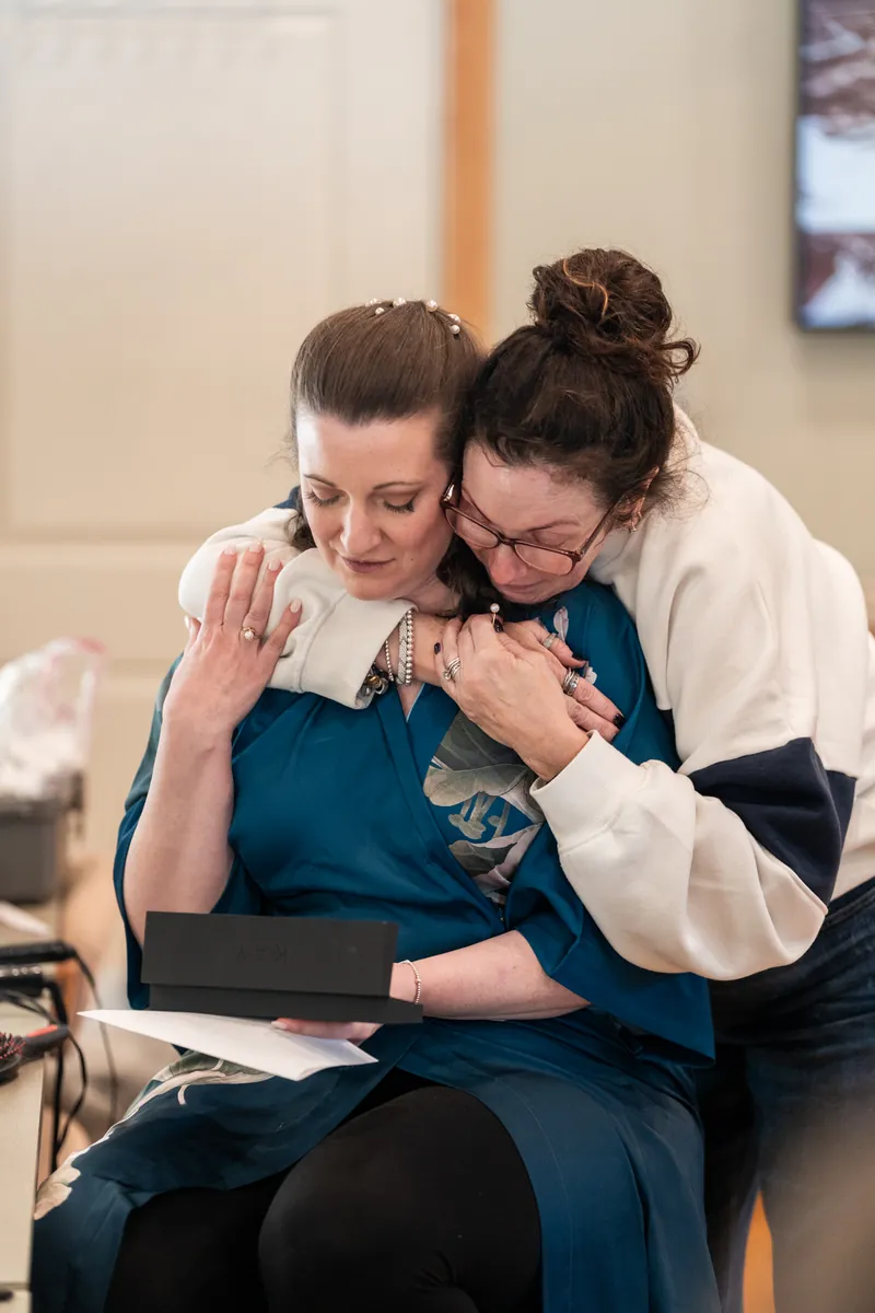 The bride in a teal floral robe sharing an emotional hug with a loved one while holding a gift box and letter