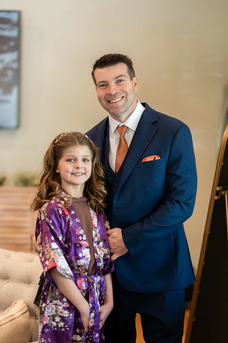 The groom in his navy suit posing with his daughter in a purple floral robe before the ceremony