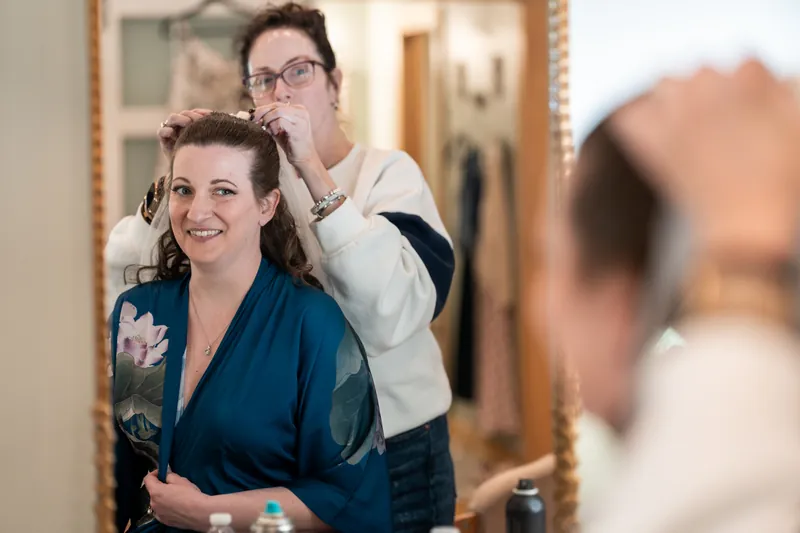 The bride smiling as her hairstylist pins her hair, reflected in a gold-framed mirror