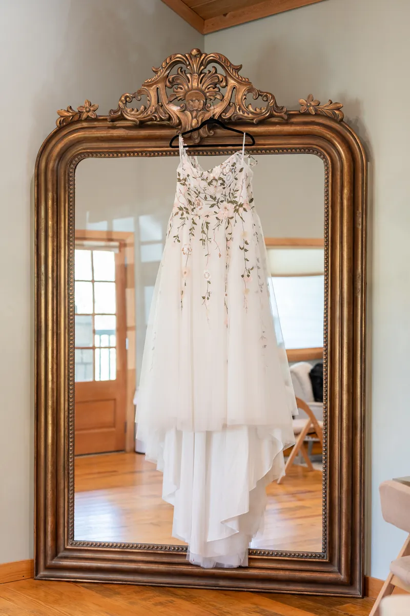 The bride's floral embroidered wedding dress hanging on an ornate gold mirror in the Fountain of Youth bridal suite