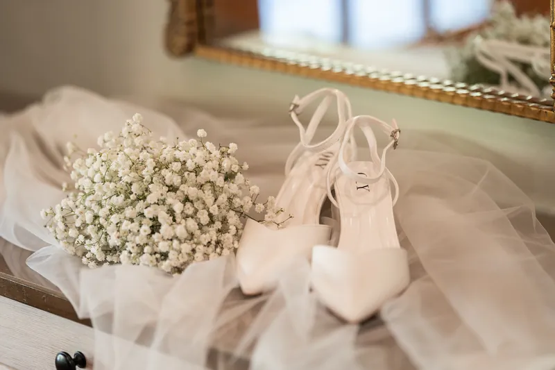 Baby's breath bouquet and white heels arranged on tulle veil beside a gold mirror