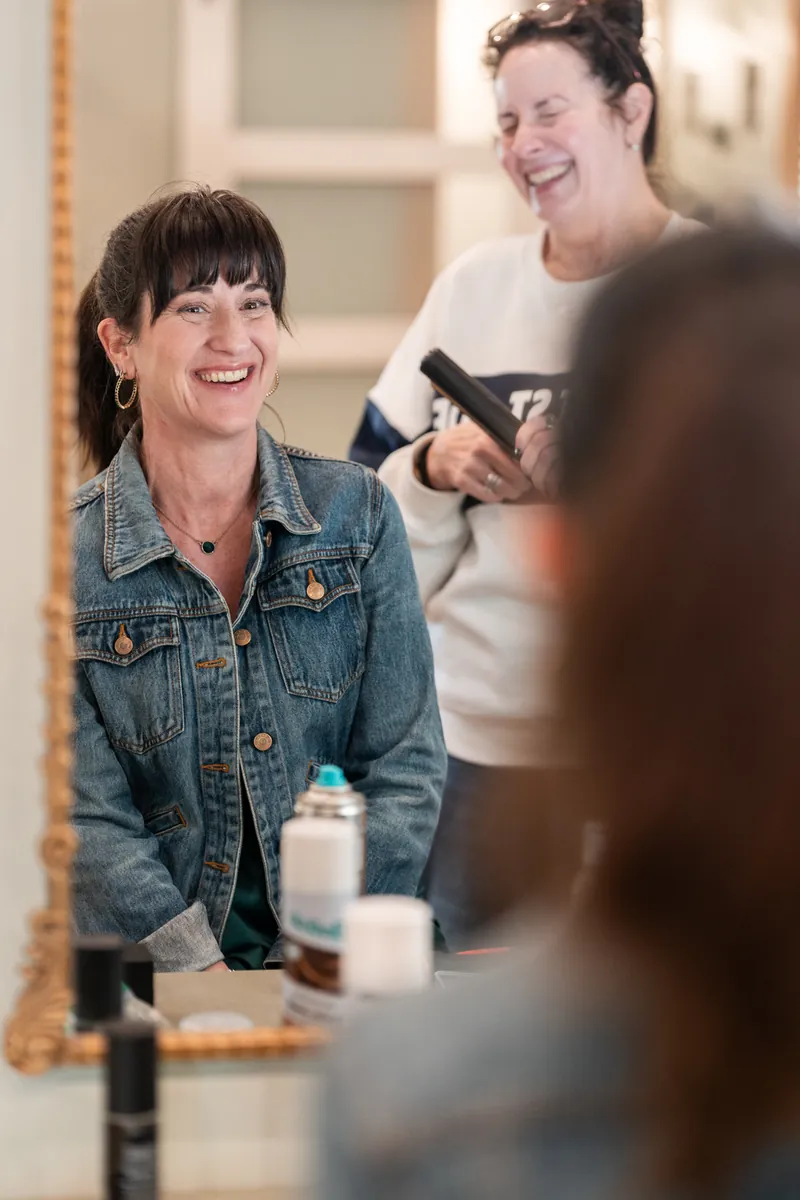 A bridesmaid in a denim jacket laughing in the mirror while the hairstylist works behind her