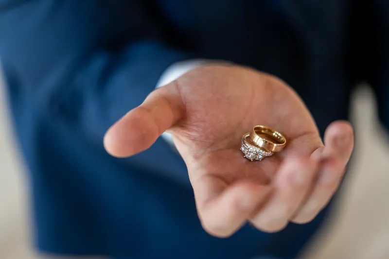 The groom's hand holding two gold wedding bands, a diamond ring and a plain gold band