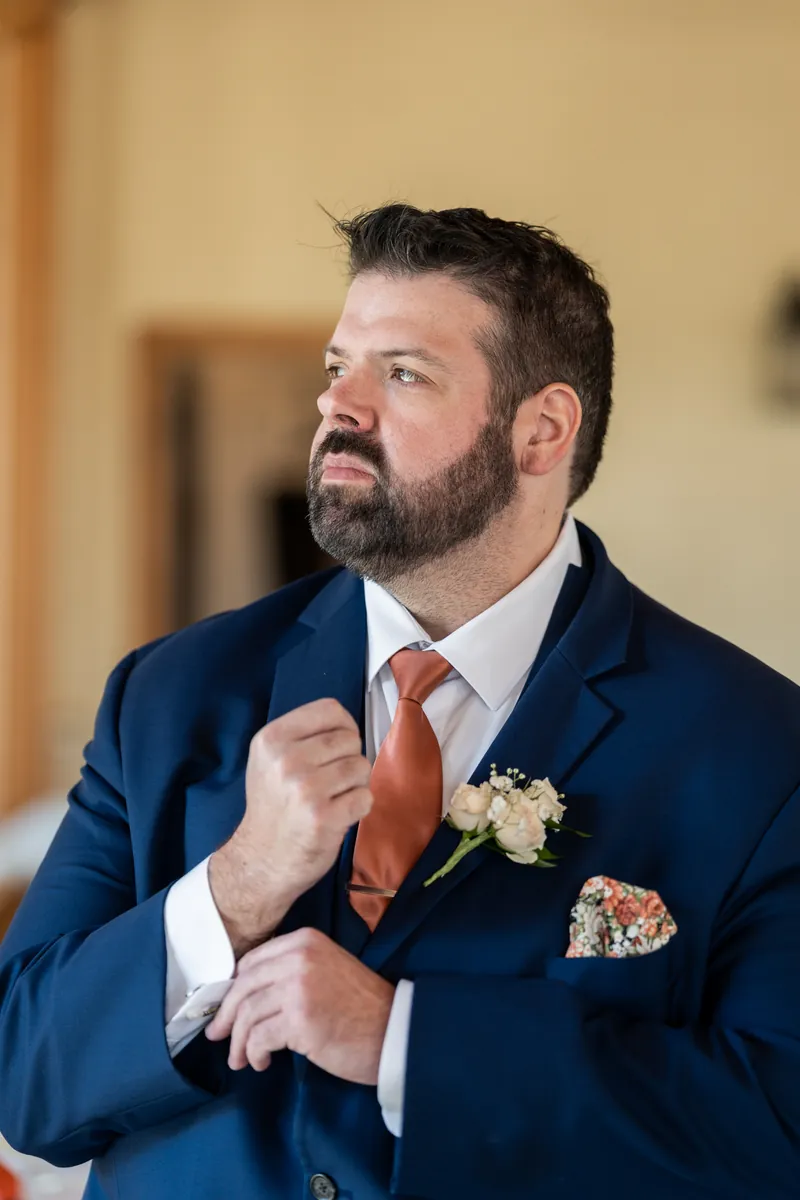 The groom adjusting his cuff in his navy suit with a rust tie, white boutonniere, and floral pocket square