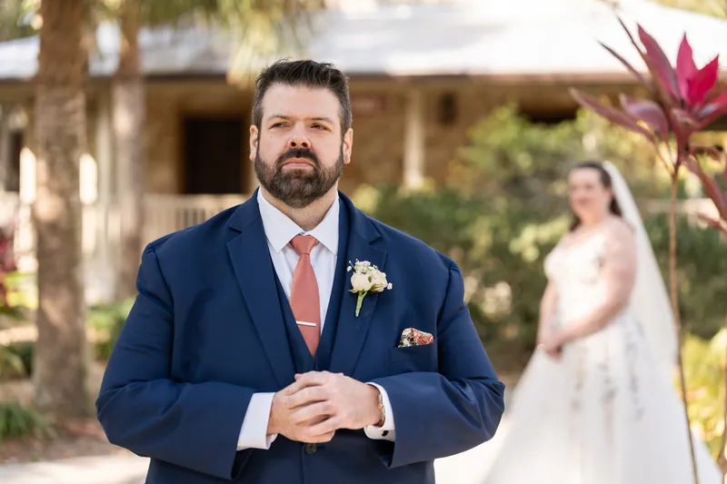 The groom waiting with hands clasped as the bride approaches from behind for their first look among tropical plants