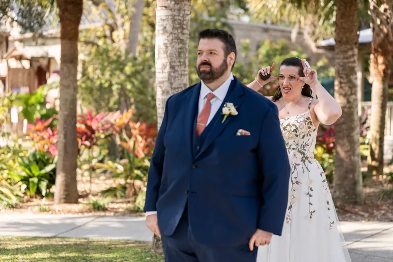 The bride sneaking up behind the groom for their first look, grinning with her hand raised, surrounded by palm trees