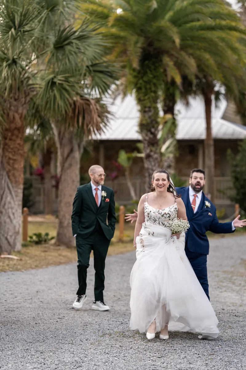 The bride laughing as she walks ahead of the best man and groom on a gravel path with palm trees behind them