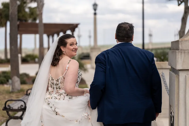 The bride glancing back over her shoulder with a smile as she and Chase walk toward the bayfront pavilion