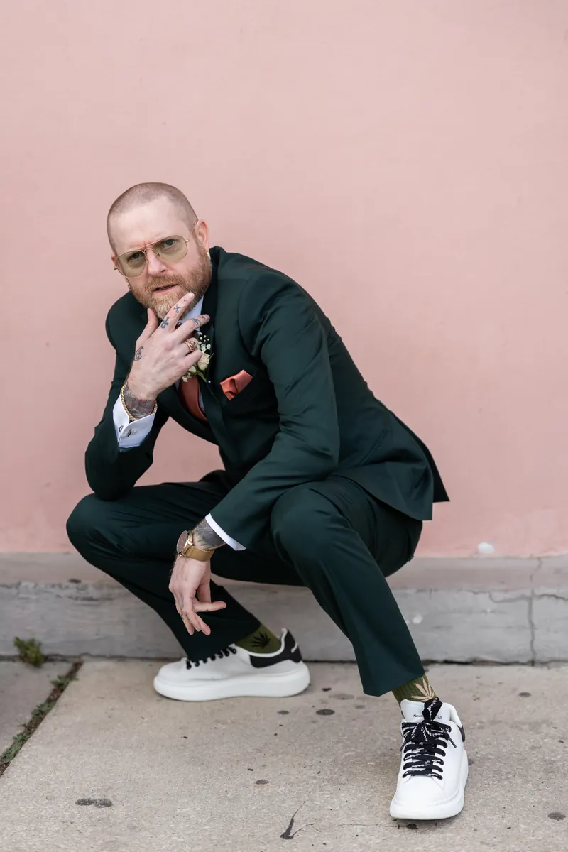 The best man in his green suit crouching against a pink wall, showing off his tattoos and sneakers