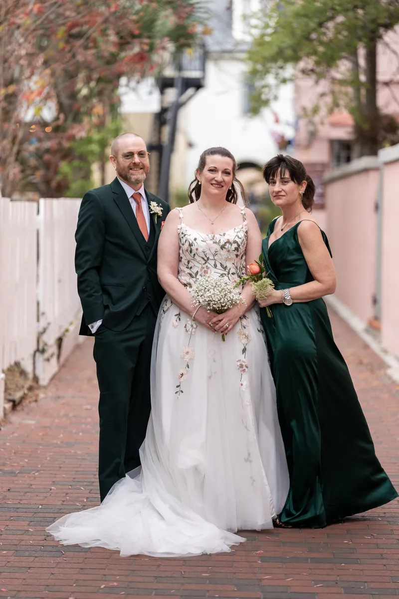 The bride with her best man and maid of honor standing together on a brick sidewalk lined with pastel buildings