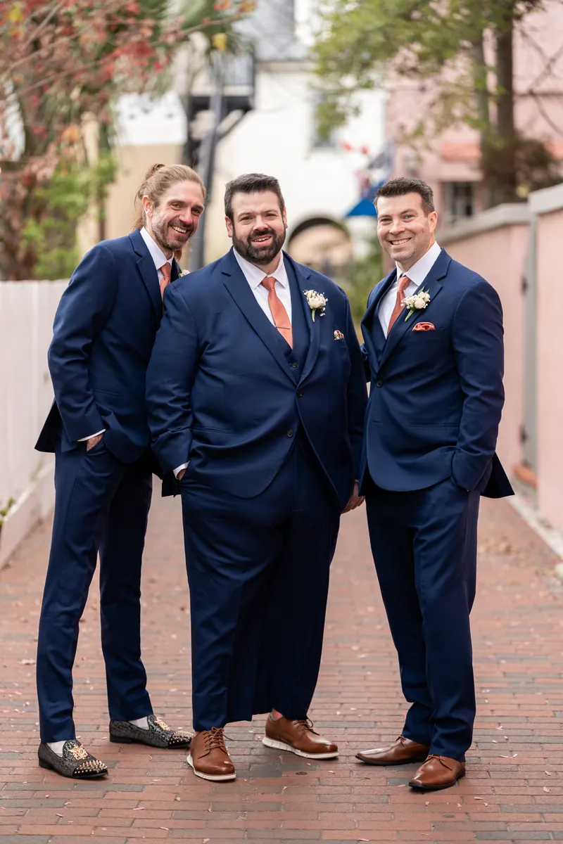 The groom with two of his groomsmen standing close together in a brick alley, all in navy suits with rust ties