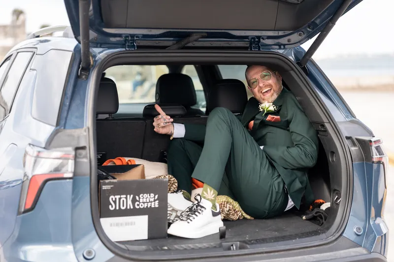The best man sitting in the back of an SUV laughing, surrounded by shoes and cold brew coffee